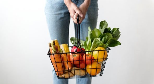 Person in jeans, visible from the waist down, holds a grocery basket in front of them filled with carrots, tomatoes, potatoes and other vegetables.