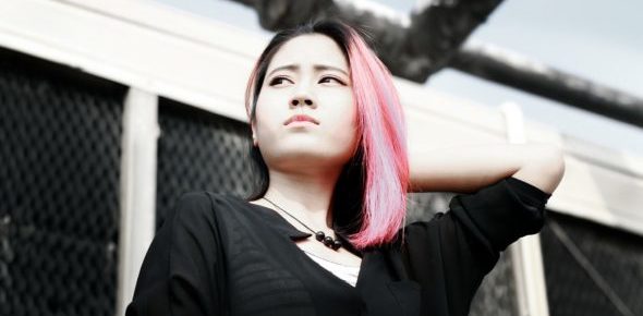 Girl of Asian heritage with pink hair looks into the distance, looking thoughtful with one hand on the back of her neck. She is outside on a sunny day.